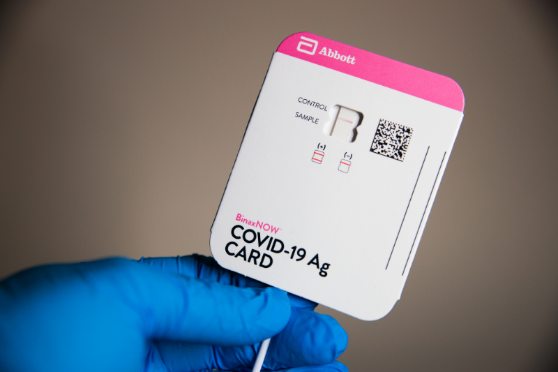 blue gloved hand holding a BinaxNow COVID-19 rapid test with a single pink line indicating a negative test